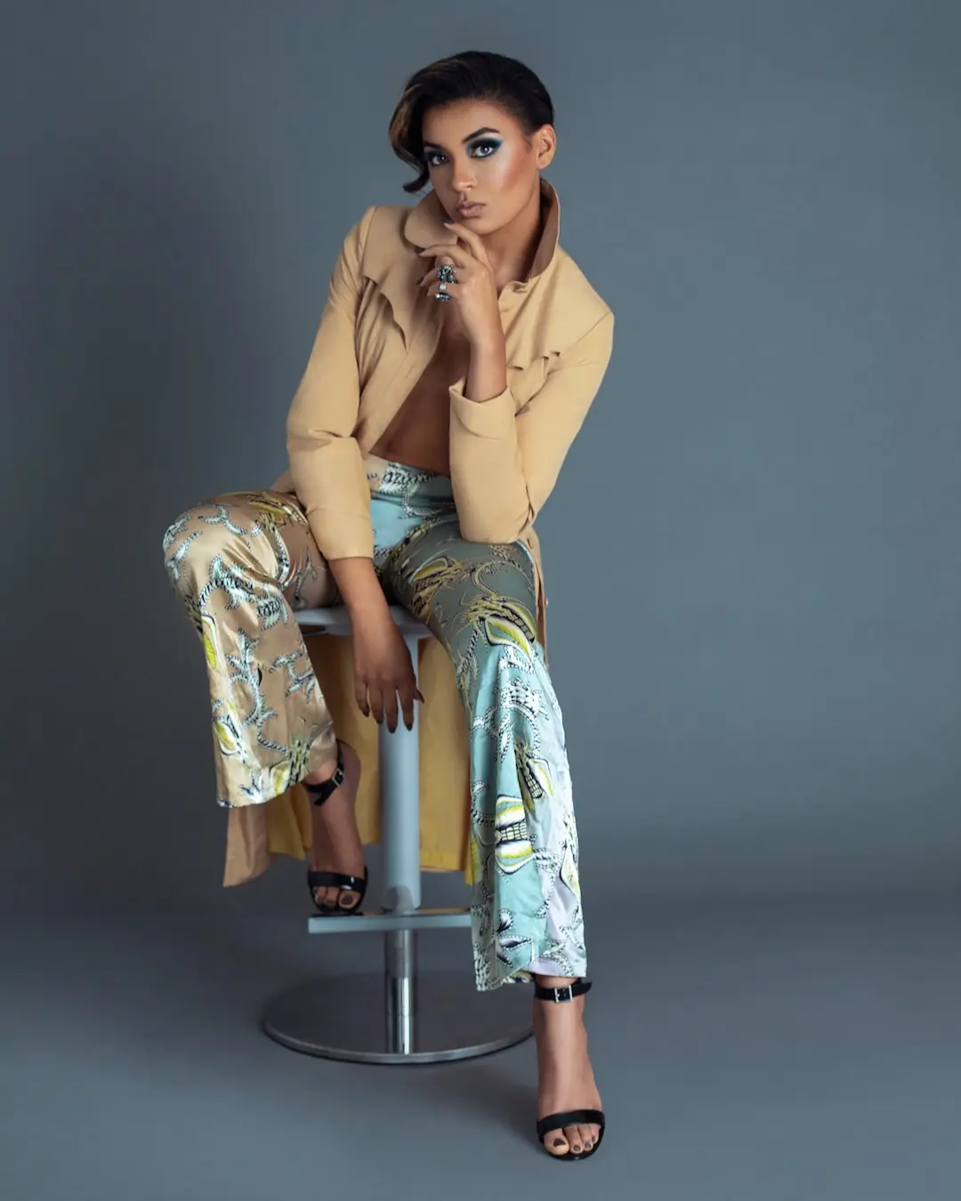 Fianna seated in a neutral coat and patterned trousers in a studio fashion portrait.