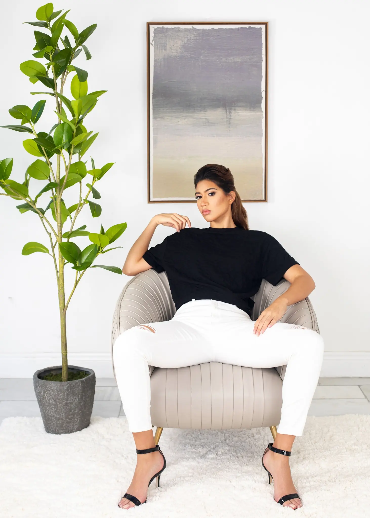 Sangella seated in a grey chair wearing a black top, white trousers and black heels in a bright fashion editorial interior
